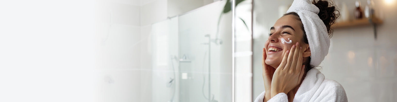 Eine Frau steht Bademantel und Handtuch um die Haare im Badezimmer, Creme an der Wange und fasst sich ins Gesicht.