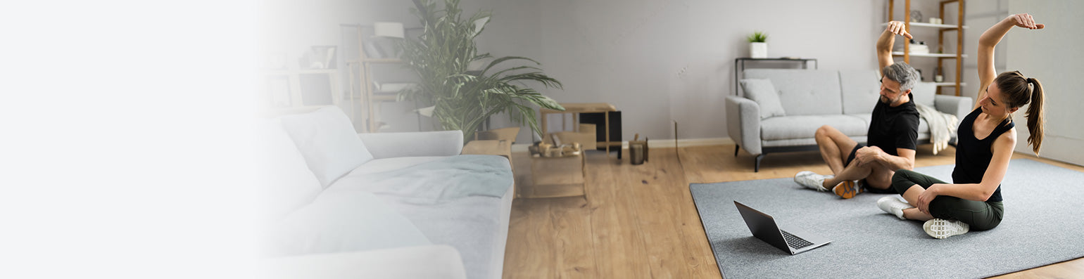 Eine Frau und ein Mann sitzen im Schneidersitz auf dem Boden vor einem Laptop in einem Wohnzimmer und machen Yoga.
