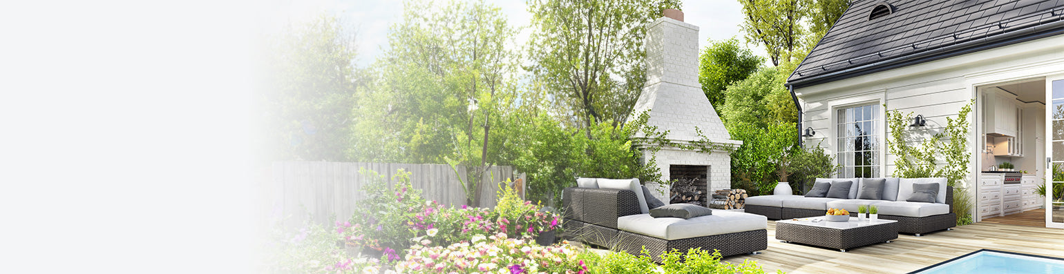 Eine moderne Terrasse mit grau-weißen Gartenmöbeln, im Hintergrund ein großer weiß gemauerter Außenkamin neben einem weißen Wohnhaus.