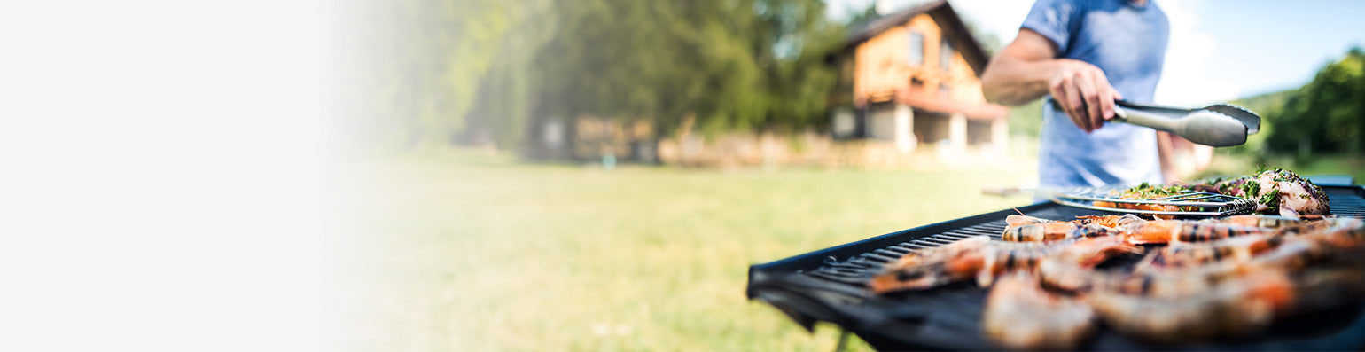Ein Mann steht mit Grillzange in der Hand an einem gut gefüllten Grillrost, im Hintergrund ein weitläufiger Garten mit Haus.