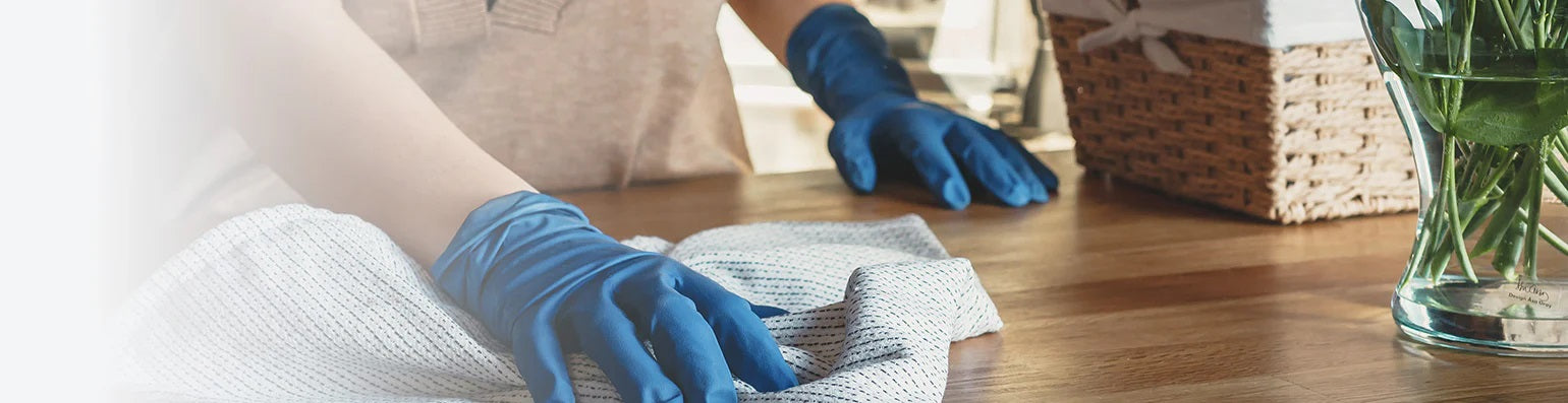 Eine Frau mit blauen Gummihandschuhen wischt mit einem grauen Tuch einen Tisch.