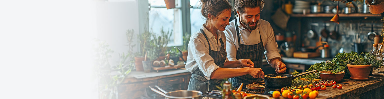 Eine Frau und einen Mann stehen gemeinam am Herd und kochen in einer rustikalen Küche mit viel Gemüse und Pflanzen.