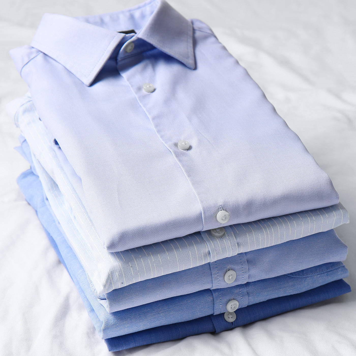 Ordentlich gefaltete, hellblaue Hemden in verschiedenen Blautönen liegen gestapelt auf einem weißen Bettlaken.